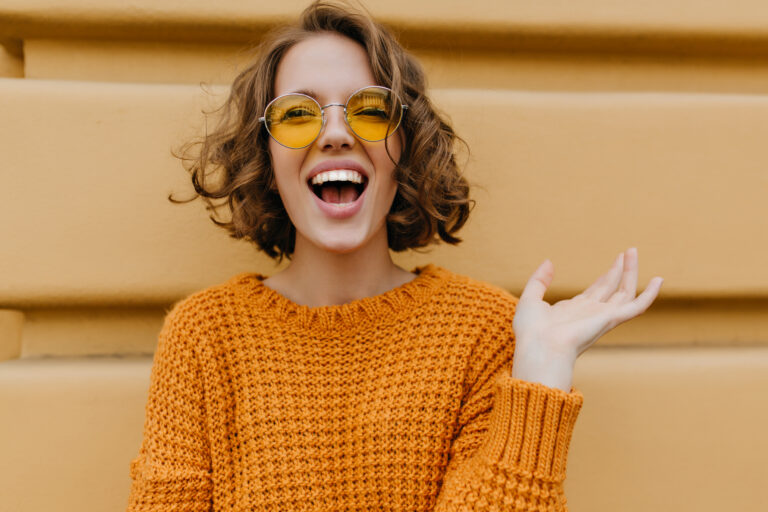 Enthusiastic smiling girl with shiny curls posing in front of old wall. Close-up outdoor portrait of enchanting lady in sweater and trendy glasses.