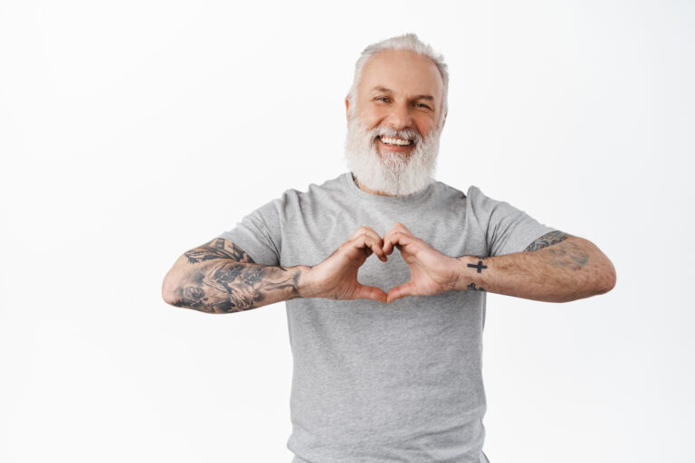 Happy mature man with tattoos laughing, showing heart I love you gesture, express sympathy, like or care for someone, standing in grey t-shirt against white background.