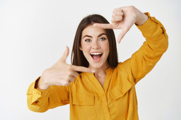 Happy young woman capturing moment, making hand frames camera gesture and smiling, standing over white background. Copy space
