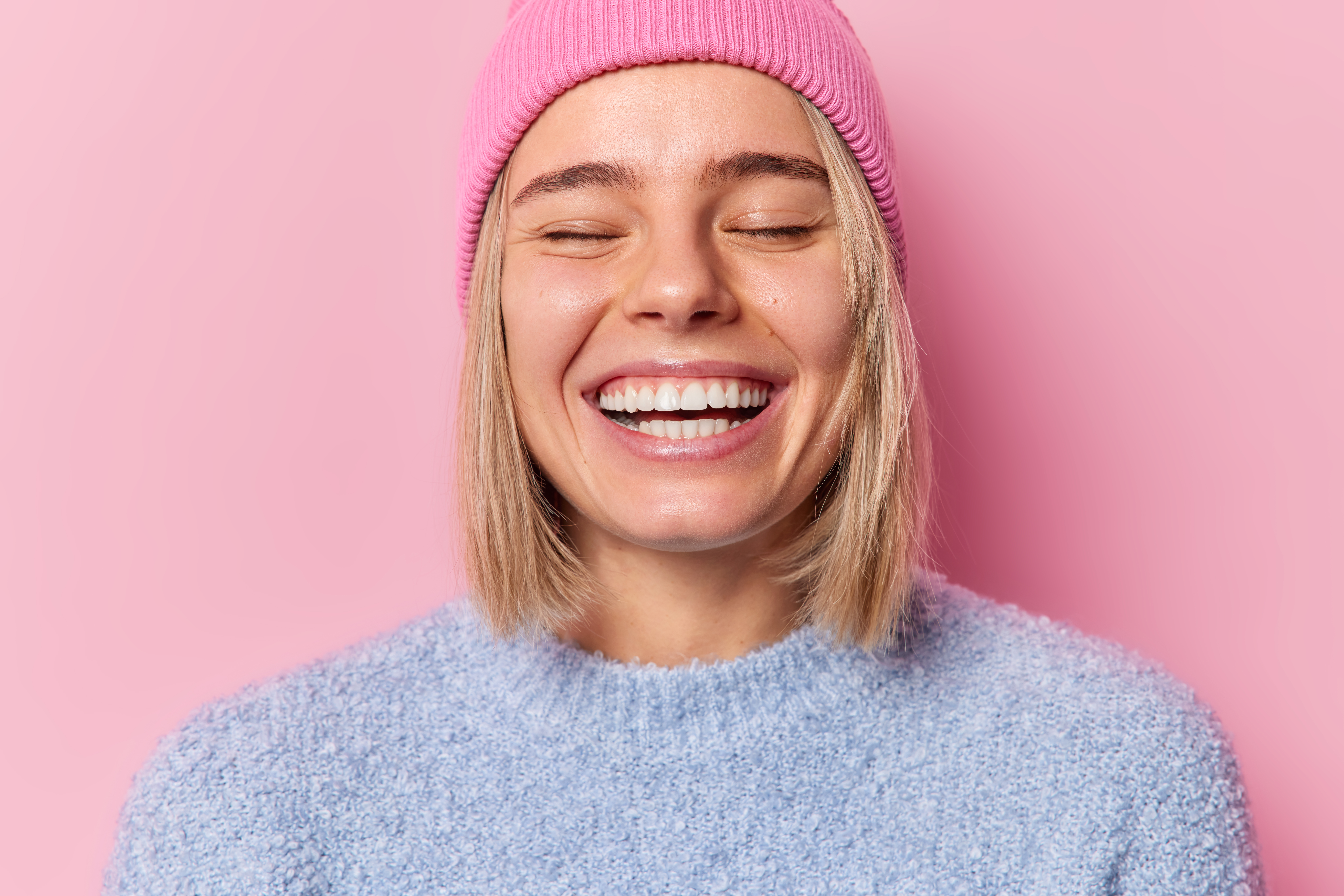 Headshot of optimistic woman keep eyes closed smile broadly show white teeth expresses sincere feelings feels very glad wears hat and casual jumper isolated over pink background. Emotions concept