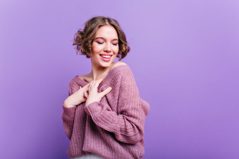 Inspired white woman wears soft knitted sweater posing with shy smile. Indoor portrait of enthusiastic young lady with trendy hairstyle laughing on purple background.