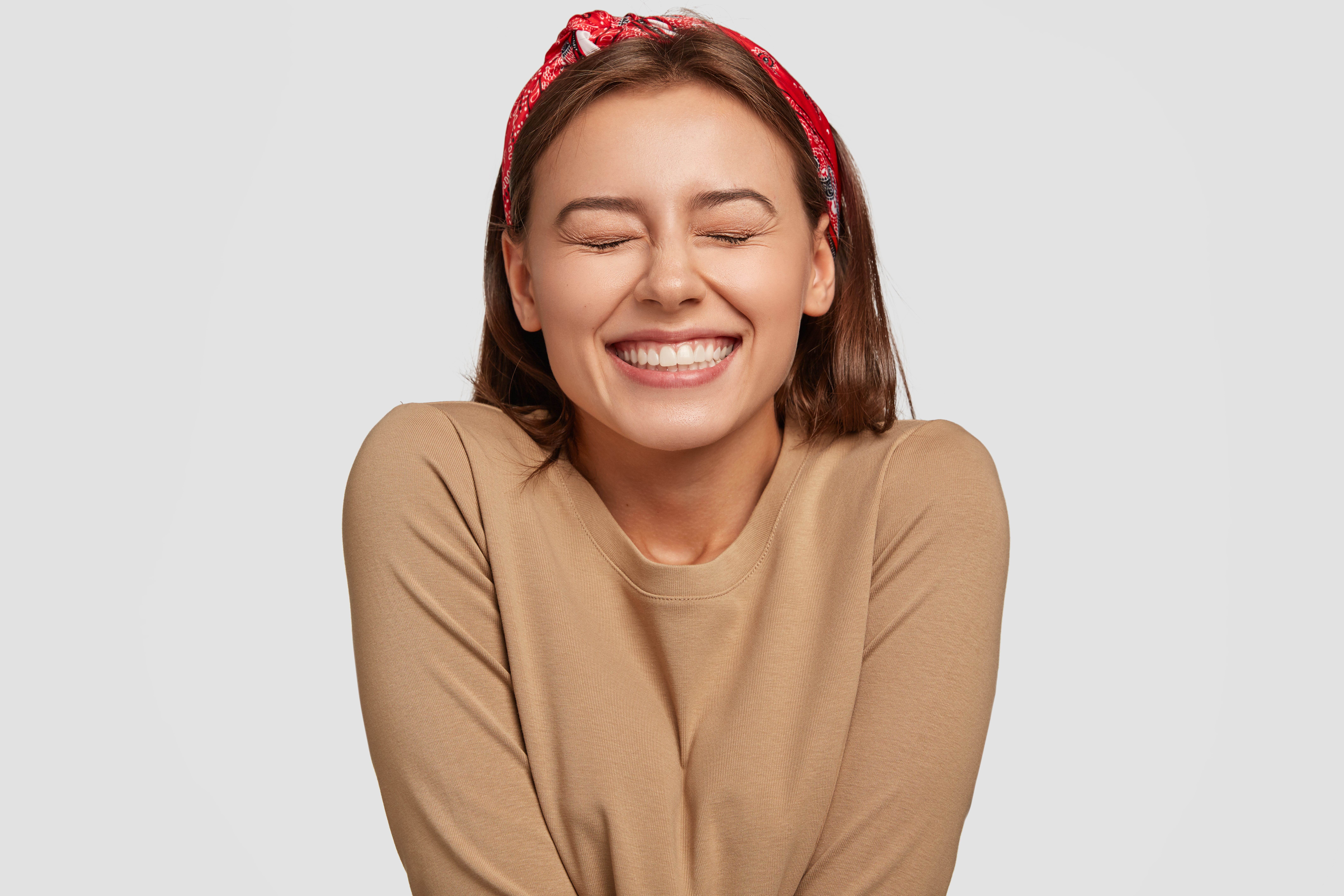 Portrait of happy European woman has broad smile, closes eyes, feels excitement, being in high spirit, recieves proposal from boyfriend, laughs at something funny, models against white background